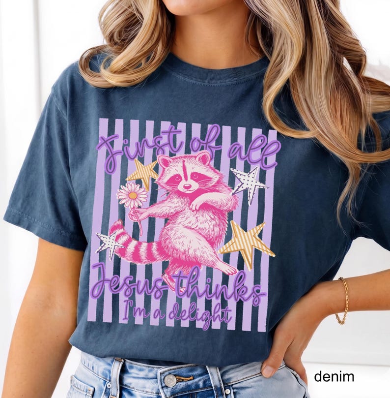 May include: Denim blue t-shirt featuring a pink raccoon holding a daisy, with the text "First of all Jesus thinks I'm a delight" in a purple and white striped design. The design includes gold star accents.