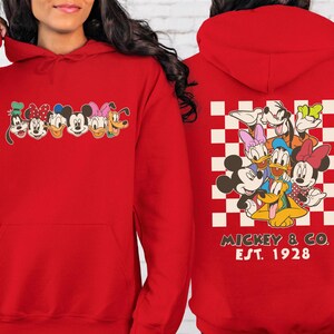 Puede incluir: Sudadera con capucha roja con un gr&aacute;fico de personajes de Disney en la parte delantera y trasera. La parte delantera muestra a Goofy, Minnie, Mickey, Daisy y Pluto. La parte trasera tiene un patr&oacute;n de cuadros con los personajes y el texto "Mickey & Co. Est. 1928."