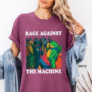 May include: Berry-colored t-shirt with the text "RAGE AGAINST THE MACHINE" in white. The graphic depicts a frog facing a group of people in riot gear. The frog is green with yellow accents and a blue scarf.