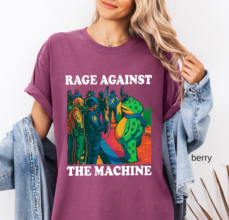 May include: Berry-colored t-shirt with the text "RAGE AGAINST THE MACHINE" in white. The graphic depicts a frog facing a group of people in riot gear. The frog is green with yellow accents and a blue scarf.