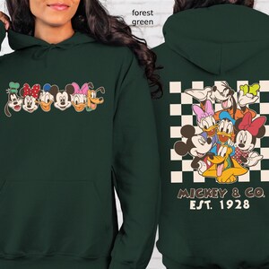 Puede incluir: Sudadera con capucha verde bosque con un gr&aacute;fico de dibujos animados de Mickey Mouse, Minnie Mouse, Donald Duck, Daisy Duck, Goofy y Pluto. La parte trasera tiene un patr&oacute;n de cuadros con los personajes y el texto "Mickey & Co. Est. 1928".