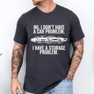May include: Dark gray t-shirt with white text and car graphics. The text reads, "NO, I DON'T HAVE A CAR PROBLEM. I HAVE A STORAGE PROBLEM." The shirt features a crew neck and short sleeves.