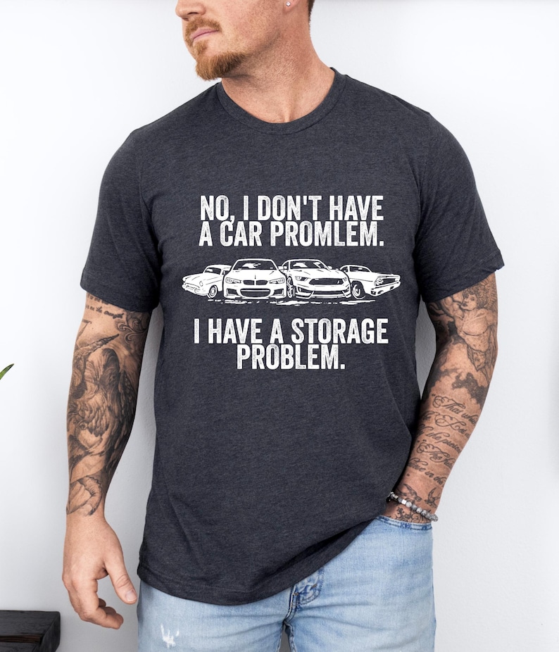 May include: Dark gray t-shirt with white text and car graphics. The text reads, "NO, I DON'T HAVE A CAR PROBLEM. I HAVE A STORAGE PROBLEM." The shirt features a crew neck and short sleeves.