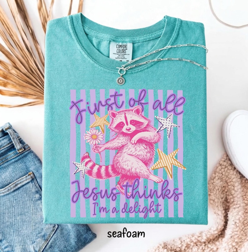 May include: Seafoam green t-shirt with a raccoon graphic holding a flower, surrounded by stars and vertical stripes. Text reads "First of all Jesus thinks I'm a delight." A silver necklace is draped on the shirt.