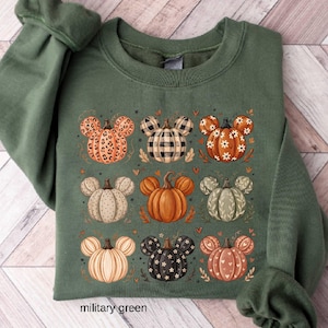 May include: A military green crewneck sweatshirt featuring a graphic print of nine pumpkins arranged in a grid. Each pumpkin has mouse-ear shapes and displays unique patterns including leopard print, black and white plaid, floral, and polka dots.