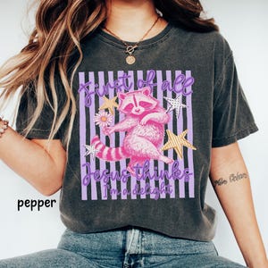 May include: Dark grey t-shirt featuring a pink raccoon holding a flower, with the text "First of all Jesus thinks I'm a delight" in purple. The design includes gold stars and a striped background.
