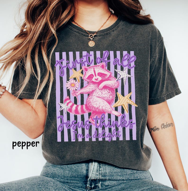 May include: Dark grey t-shirt featuring a pink raccoon holding a flower, with the text "First of all Jesus thinks I'm a delight" in purple. The design includes gold stars and a striped background.