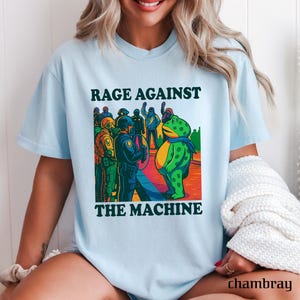May include: Light blue t-shirt with the text "RAGE AGAINST THE MACHINE" above a cartoon illustration. The illustration depicts a green frog facing a group of police officers. The frog wears a blue scarf. The shirt is made of chambray material.