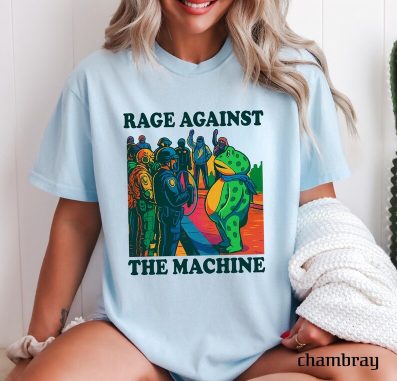 May include: Light blue t-shirt with the text "RAGE AGAINST THE MACHINE" above a cartoon illustration. The illustration depicts a green frog facing a group of police officers. The frog wears a blue scarf. The shirt is made of chambray material.