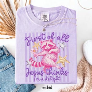 May include: Orchid-colored t-shirt with a graphic of a pink raccoon holding a daisy, surrounded by stars. Text reads "First of all Jesus thinks I'm a delight." A silver necklace with a sun pendant is on the shirt.