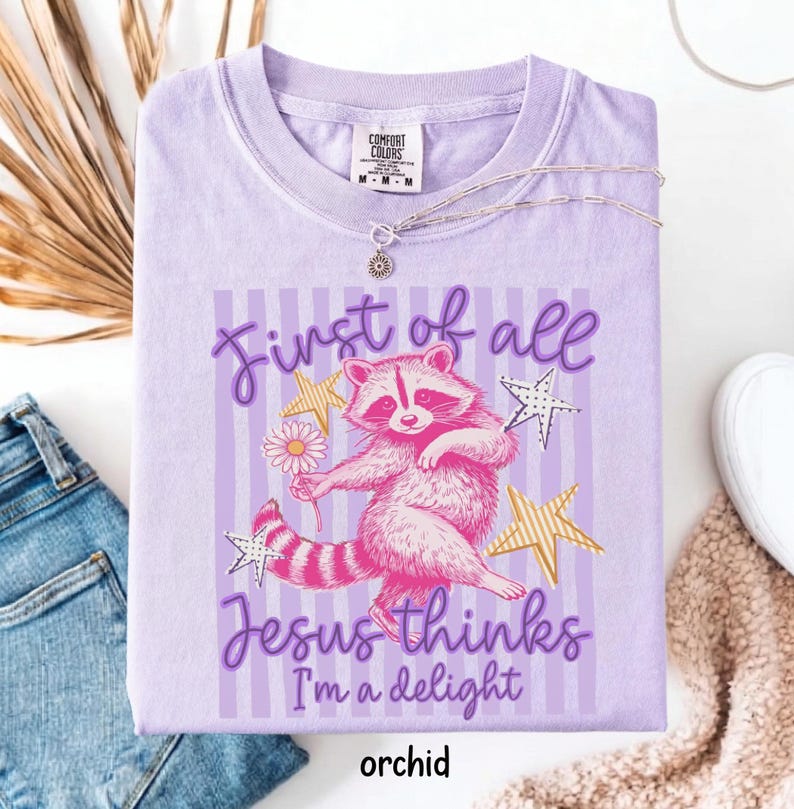 May include: Orchid-colored t-shirt with a graphic of a pink raccoon holding a daisy, surrounded by stars. Text reads "First of all Jesus thinks I'm a delight." A silver necklace with a sun pendant is on the shirt.