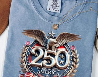 May include: A blue jean Comfort Colors t-shirt featuring a graphic commemorating the 250th anniversary of the United States of America. The design includes an eagle, stars, and the years 1776-2026. A gold necklace with a pendant rests on the shirt.