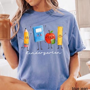 May include: A blue t-shirt featuring a graphic of a yellow pencil, a blue notebook, a red apple with glasses, and a yellow ruler, all with smiling faces and limbs. The word Kindergarten is printed in white cursive script below the characters.