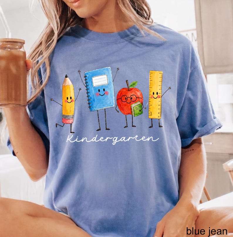 May include: A blue t-shirt featuring a graphic of a yellow pencil, a blue notebook, a red apple with glasses, and a yellow ruler, all with smiling faces and limbs. The word Kindergarten is printed in white cursive script below the characters.