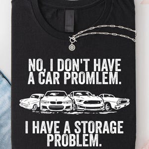 May include: Black t-shirt with the text "NO, I DON'T HAVE A CAR PROBLEM. I HAVE A STORAGE PROBLEM." and illustrations of four cars. A silver necklace with a pendant is draped across the shirt.