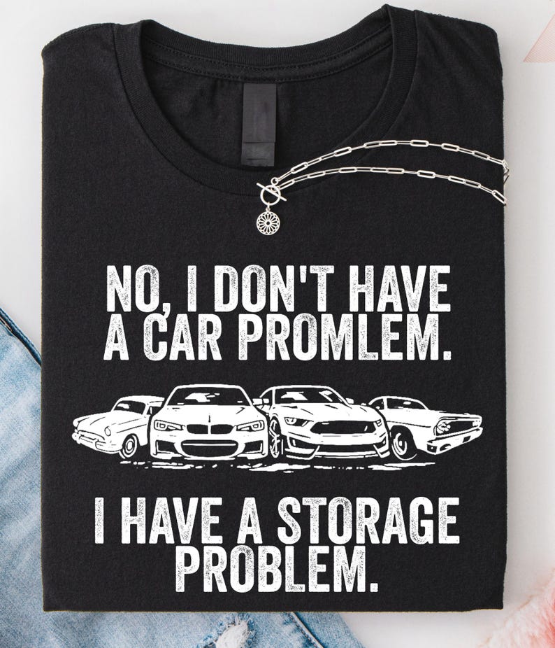 May include: Black t-shirt with the text "NO, I DON'T HAVE A CAR PROBLEM. I HAVE A STORAGE PROBLEM." and illustrations of four cars. A silver necklace with a pendant is draped across the shirt.