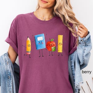 May include: A berry-colored t-shirt featuring a graphic print of four anthropomorphic school supplies: a yellow pencil, a blue spiral notebook, a red apple wearing glasses and holding a small green book, and a yellow ruler, all with smiling faces.