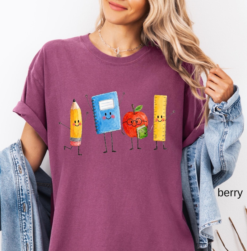 May include: A berry-colored t-shirt featuring a graphic print of four anthropomorphic school supplies: a yellow pencil, a blue spiral notebook, a red apple wearing glasses and holding a small green book, and a yellow ruler, all with smiling faces.