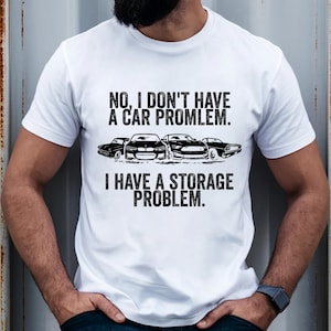May include: White t-shirt with the text "NO, I DON'T HAVE A CAR PROBLEM. I HAVE A STORAGE PROBLEM." The graphic includes black car silhouettes. The shirt is a casual wear item.