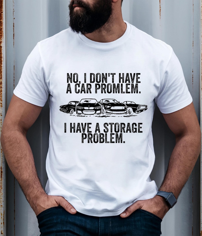 May include: White t-shirt with the text "NO, I DON'T HAVE A CAR PROBLEM. I HAVE A STORAGE PROBLEM." The graphic includes black car silhouettes. The shirt is a casual wear item.