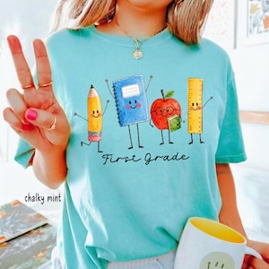 May include: A chalky mint t-shirt featuring a graphic of a yellow pencil, a blue notebook, a red apple with glasses, and a yellow ruler with arms and legs. The text "First Grade" is printed in black cursive script below the school supply characters.