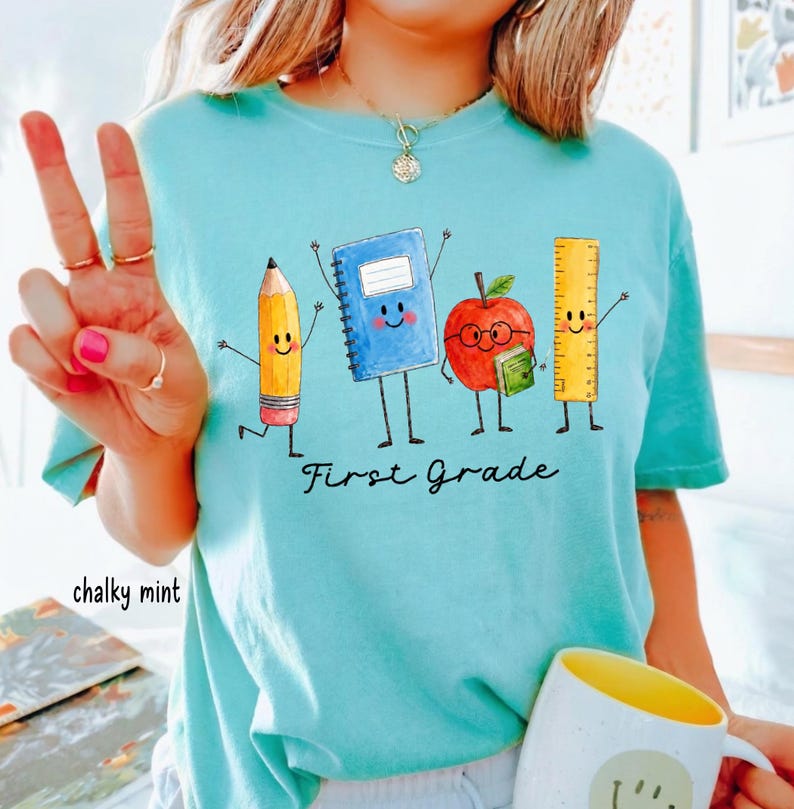May include: A chalky mint t-shirt featuring a graphic of a yellow pencil, a blue notebook, a red apple with glasses, and a yellow ruler with arms and legs. The text "First Grade" is printed in black cursive script below the school supply characters.