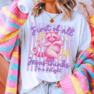 May include: White t-shirt with a pink raccoon graphic holding a flower, surrounded by gold stars. The text reads "First of all Jesus thinks I'm a delight" in purple script. The shirt is worn with a colorful striped cardigan.