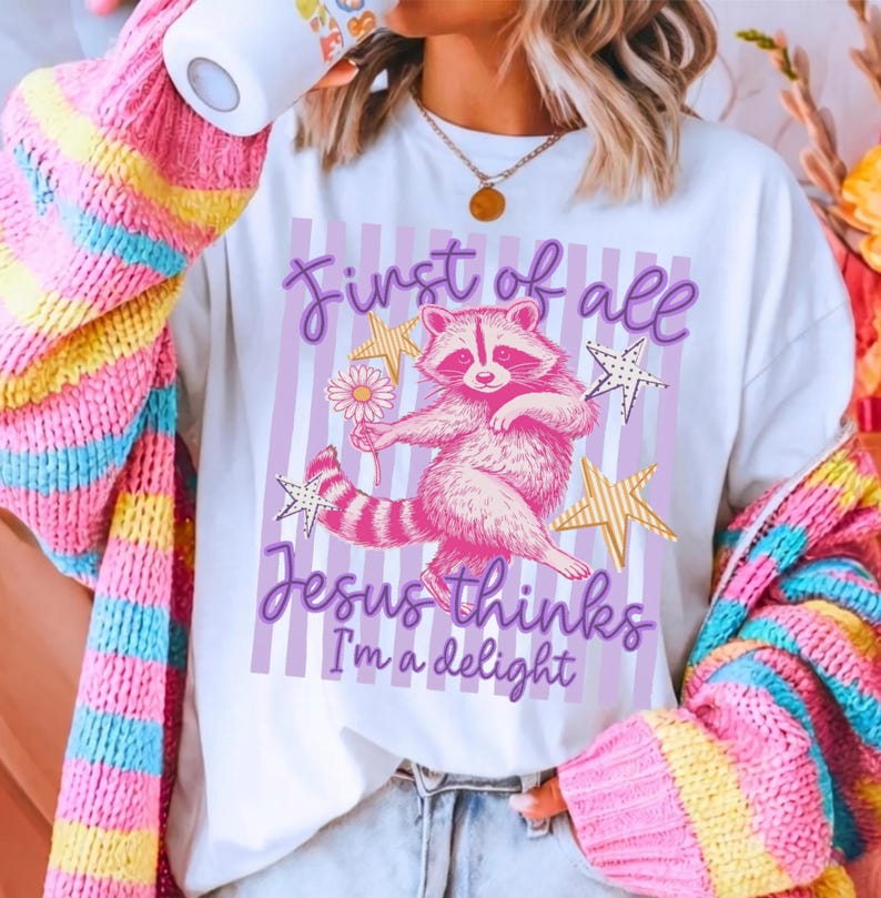 May include: White t-shirt with a pink raccoon graphic holding a flower, surrounded by gold stars. The text reads "First of all Jesus thinks I'm a delight" in purple script. The shirt is worn with a colorful striped cardigan.