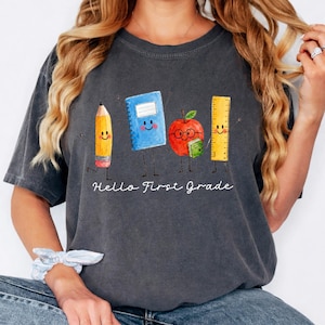 May include: A charcoal gray short-sleeve t-shirt featuring a watercolor-style graphic of a smiling pencil, a blue notebook, a red apple with glasses holding a small green book, and a yellow ruler. The text "Hello First Grade" is printed below.