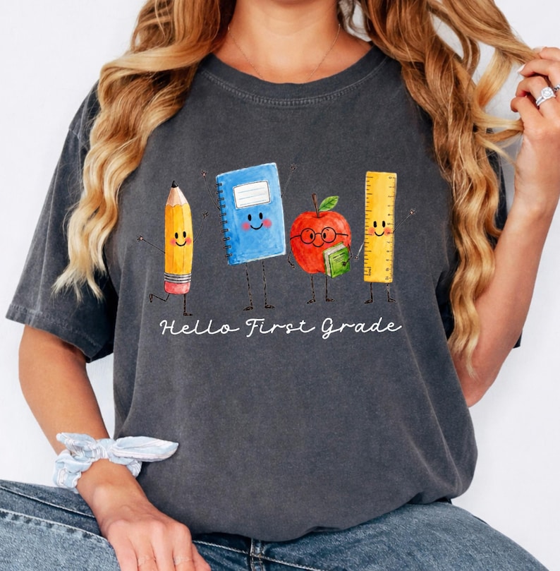 May include: A charcoal gray short-sleeve t-shirt featuring a watercolor-style graphic of a smiling pencil, a blue notebook, a red apple with glasses holding a small green book, and a yellow ruler. The text "Hello First Grade" is printed below.