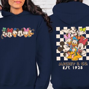 Puede incluir: Sudadera con capucha azul marino con un dise&ntilde;o de personajes de Disney en la parte delantera. La parte trasera muestra un grupo de personajes sobre un fondo a cuadros, con "Mickey & Co. Est. 1928" impreso debajo. La sudadera tiene capucha con cord&oacute;n y bolsillo delantero.