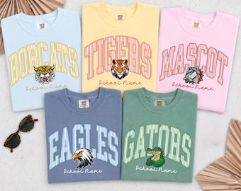 May include: Five crewneck t-shirts in light blue, yellow, pink, slate blue, and sage green. Each shirt features a collegiate-style arched word, a mascot illustration, and the text School Name. A pair of tortoise shell sunglasses is on the left.