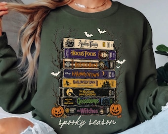 May include: A military green crewneck sweatshirt featuring a graphic of stacked VHS tapes titled The Addams Family, Hocus Pocus, Beetlejuice, Halloweentown, Halloweentown II, The Nightmare Before Christmas, Goosebumps, and The Witches, with spooky season text.