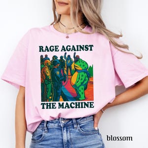 May include: Light pink t-shirt with the text "RAGE AGAINST THE MACHINE" above and below a colorful graphic. The graphic depicts a frog in a blue scarf facing a group of police officers. The word "blossom" is printed at the bottom right.