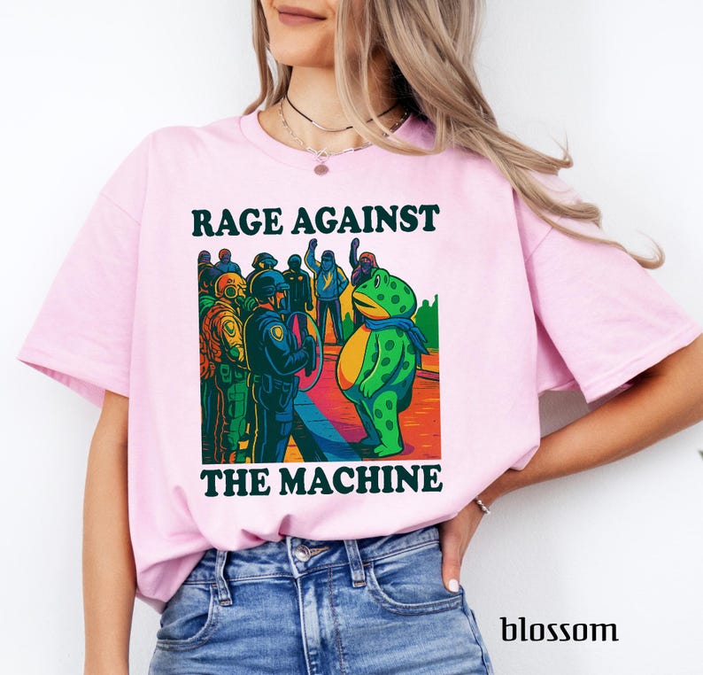 May include: Light pink t-shirt with the text "RAGE AGAINST THE MACHINE" above and below a colorful graphic. The graphic depicts a frog in a blue scarf facing a group of police officers. The word "blossom" is printed at the bottom right.