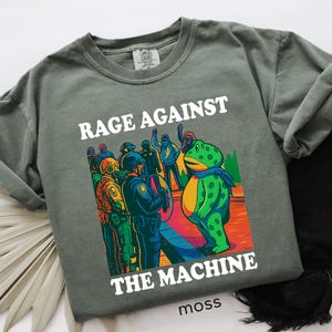 May include: Moss green t-shirt with the words "RAGE AGAINST THE MACHINE" in white. The graphic depicts a frog facing a group of people in riot gear. The word "moss" is printed at the bottom.