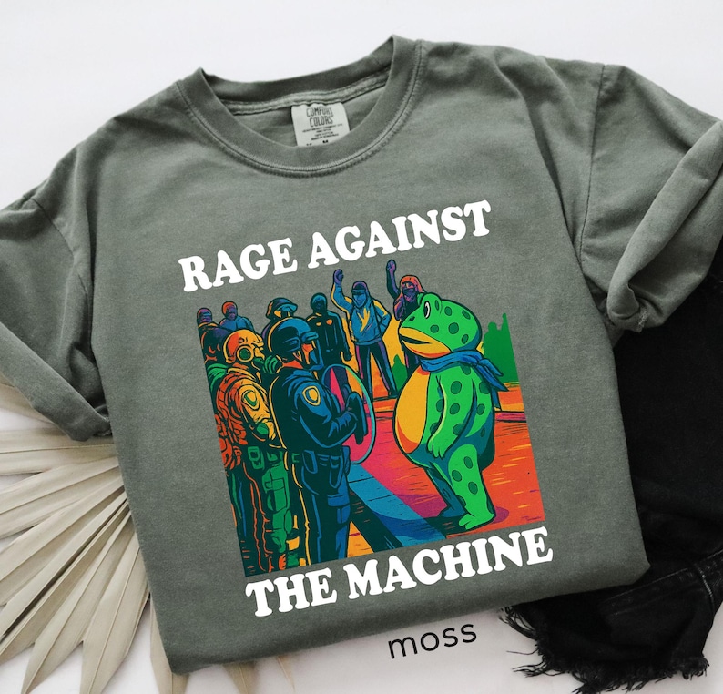 May include: Moss green t-shirt with the words "RAGE AGAINST THE MACHINE" in white. The graphic depicts a frog facing a group of people in riot gear. The word "moss" is printed at the bottom.