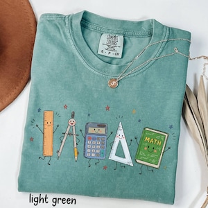 May include: A light green short-sleeve t-shirt featuring a graphic print of five animated school supplies: a ruler, a compass, a calculator, a triangle, and a green book labeled MATH. The shirt has a crew neckline and a Comfort Colors brand tag.