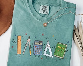 May include: A light green short-sleeve t-shirt featuring a graphic print of five animated school supplies: a ruler, a compass, a calculator, a triangle, and a green book labeled MATH. The shirt has a crew neckline and a Comfort Colors brand tag.