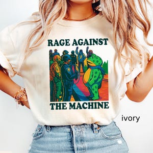 May include: Ivory-colored t-shirt with the text "RAGE AGAINST THE MACHINE" above a graphic of a frog facing a group of people. The frog wears a blue scarf. The graphic is in shades of green, blue, yellow, and red.
