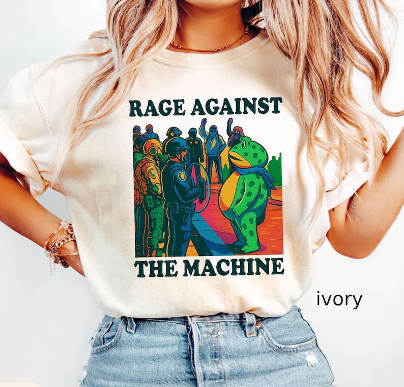 May include: Ivory-colored t-shirt with the text "RAGE AGAINST THE MACHINE" above a graphic of a frog facing a group of people. The frog wears a blue scarf. The graphic is in shades of green, blue, yellow, and red.