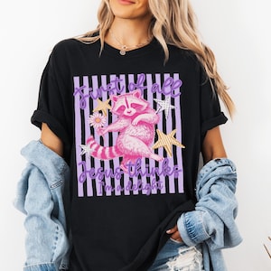 May include: Black t-shirt featuring a pink raccoon graphic with the text "First of all Jesus thinks I'm a delight." The raccoon is holding a flower and surrounded by stars. The design is set against a striped background.