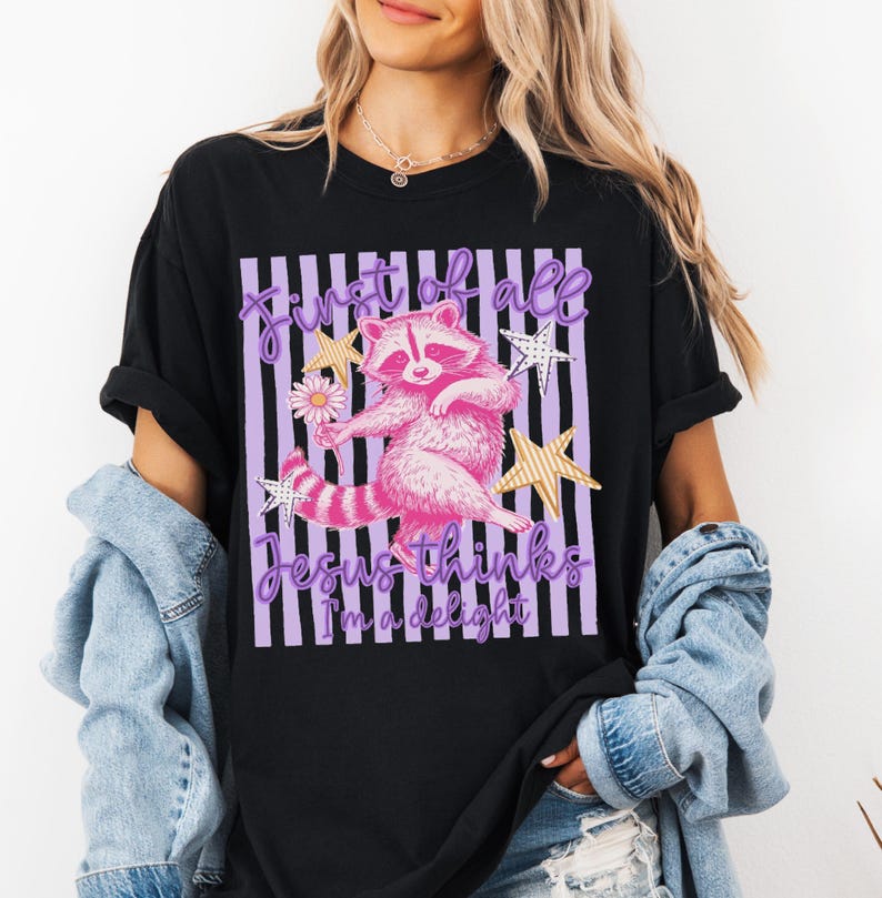 May include: Black t-shirt featuring a pink raccoon graphic with the text "First of all Jesus thinks I'm a delight." The raccoon is holding a flower and surrounded by stars. The design is set against a striped background.