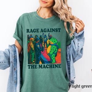 May include: Light green t-shirt with the text "RAGE AGAINST THE MACHINE" in black. The graphic features a cartoon frog and a group of people facing police officers. The shirt is a solid color and has rolled-up sleeves.