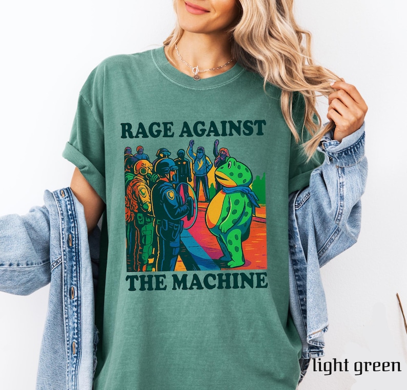 May include: Light green t-shirt with the text "RAGE AGAINST THE MACHINE" in black. The graphic features a cartoon frog and a group of people facing police officers. The shirt is a solid color and has rolled-up sleeves.