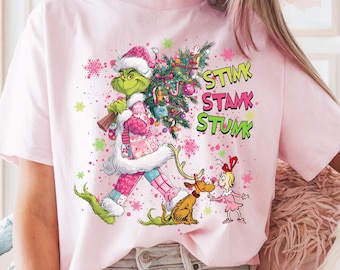 Puede incluir: Camiseta rosa claro con un diseño festivo de un personaje verde llevando un árbol de Navidad, acompañado del texto "STIMK STAMK STUMK". El diseño incluye un reno y una niña con un vestido rosa. La camiseta tiene cuello redondo y mangas cortas.