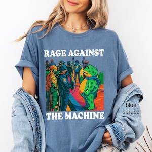 May include: Blue spruce t-shirt with the text "RAGE AGAINST THE MACHINE" above a colorful graphic. The graphic depicts a frog facing a group of police officers. The frog is wearing a blue scarf.
