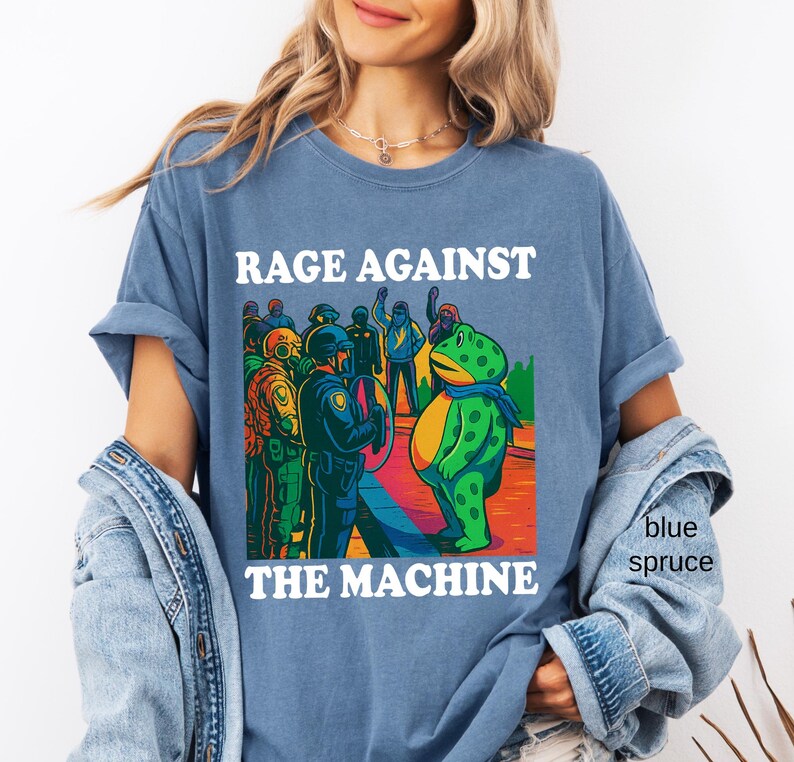 May include: Blue spruce t-shirt with the text "RAGE AGAINST THE MACHINE" above a colorful graphic. The graphic depicts a frog facing a group of police officers. The frog is wearing a blue scarf.