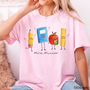 May include: A light pink t-shirt featuring a graphic illustration of four anthropomorphic school supplies: a yellow pencil, a blue notebook, a red apple with glasses holding a green book, and a yellow ruler. The text Mrs. Miller is printed below.