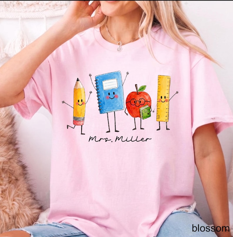May include: A light pink t-shirt featuring a graphic illustration of four anthropomorphic school supplies: a yellow pencil, a blue notebook, a red apple with glasses holding a green book, and a yellow ruler. The text Mrs. Miller is printed below.